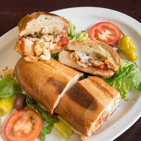 A chicken parmesan sub, cut in half, with lettuce, tomatoes, and peppers on a white plate.