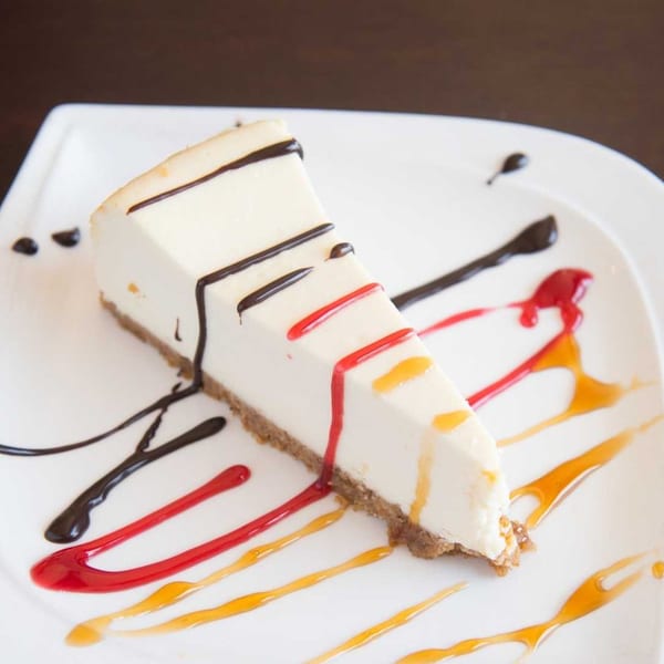 A slice of cheesecake with chocolate, red, and orange drizzles on a white plate.